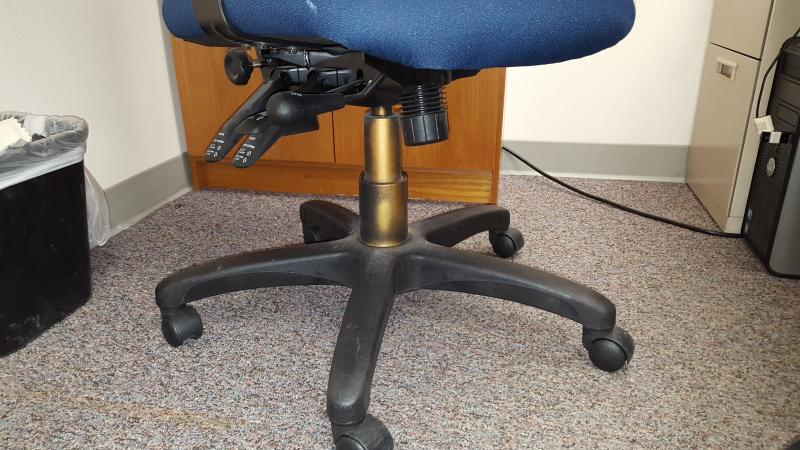 Image of a the bottom section of a chair. Underneath the seat there is a support beam connecting the seat of the chair with the legs and wheels. This support beam is normally black, but in this picture it is spray painted gold to indicate that it is a chair used for students with disabilities and with a furniture accommodations.
