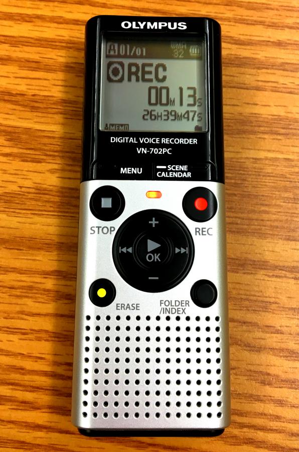 Image of a audio recorder. The brand is Olympus, it is a digital voice recorder, and the model number is VN-702PC. There is a screen displaying the time of the recording. Under the screen is a record and stop button and small speaker. The device is about one inch wide, 5 inches tall, and half an inch thick.