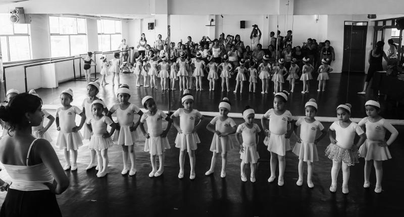 black and white photo of a line of young ballet dancers with a reflection in the mirror of the parents and guardians taking pictures and watching