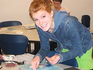 Girl with red hair and wearing a jean jacket smiles at the camera while working on a craft project